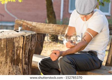 Addicted man sitting on a bench outdoors injecting his left arm intravenously