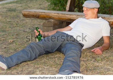 Drunk man fallen down on the ground with a bottle in his hand leaning his head on a bench in the park