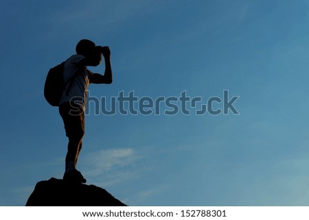 Silhouette of a male hiker looking in binoculars from the mountain top, in the evening