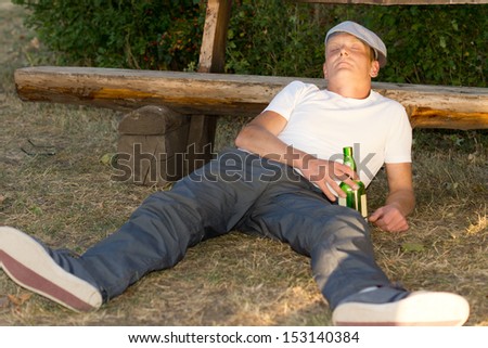 Alcoholic passed out on the ground in a park lying with his head cradled against a wooden bench holding his bottle of alcohol in his hand