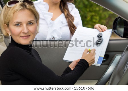 Attractive mature blond woman purchasing a new car about to sign the contract being held through the open window by the saleslady