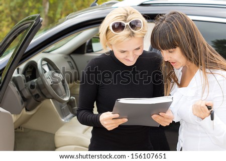 Blond woman smiling as she reads a contract for the purchase of her new car with the saleslady who is holding the ignition keys