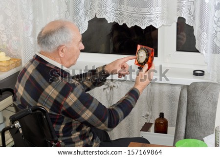 Elderly man in a wheelchair checking the clock as he sits in front of a window at night waiting for somebody to arrive