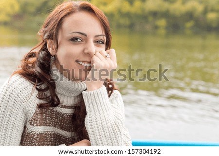 Beautiful young woman smiling happily as she sits alongside a river or lake in a warm polo-neck jersey in autumn enjoying the healthy outdoors