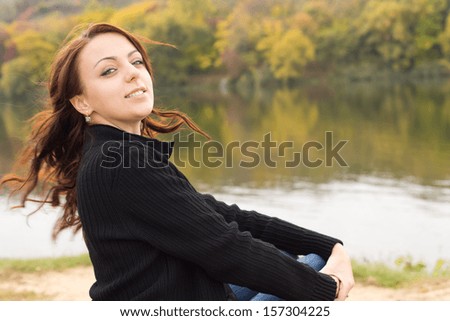 Happy playful young woman sitting above a tranquil river or lake turning back to look at the camera with her long brunette hair flying free