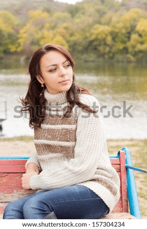 Attractive young brunette woman in a stylish polo-neck jersey relaxing at the side of an autumn lake sitting on a wooden bench looking thoughtfully off to the side