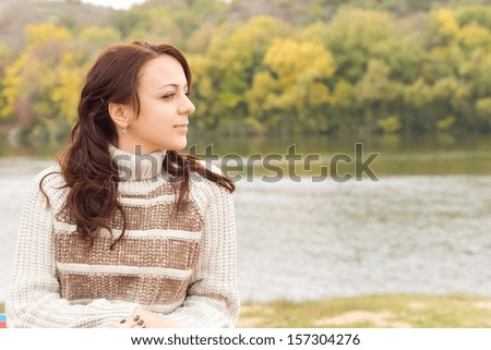 Beautiful girl enjoying the autumn weather sitting outdoors with a lovely smile on her face in a warm polo-neck sweater in front of a tranquil lake or river