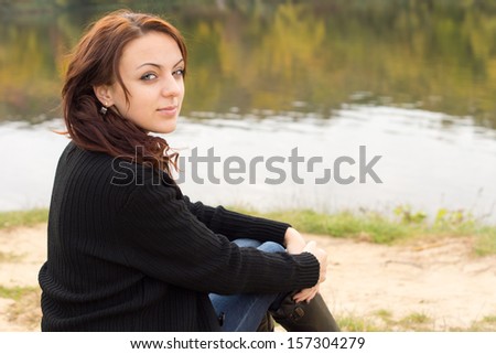 Beautiful young woman with a serene smile sitting alongside the calm water of a river or lake turning to look at the camera