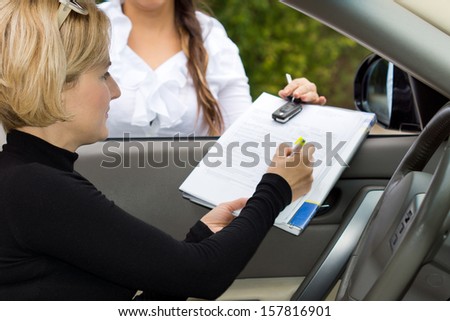 Blond female driver signing the deal on the purchase of a new car on the contact being held through the open window by the saleslady