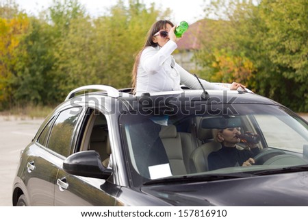 Two women drinking and driving both upending bottles to gulp down the alcohol with the passenger standing up through the sunroof