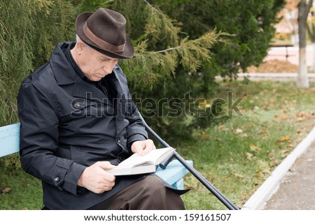 Disabled retired man on crutches reading in the park sitting on a wooden bench in a warm overcoat and hat with his crutches alongside him