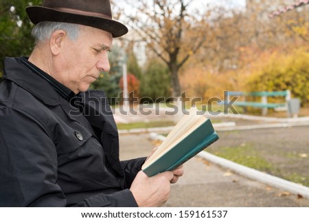 Close up side view portrait of an elderly gentleman in an overcoat and hat reading his book outdoors in the autumn sun and enjoying the peace of the public park