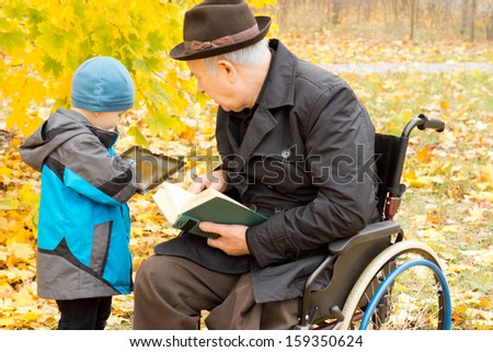 Young boy showing his grandfather something on his tablet computer as the old man sits in a wheelchair in his overcoat and hat enjoying a book in a colourful autumn garden