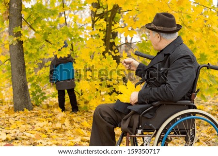 Senior disabled man with one leg amputated in a wheelchair sitting in colourful yellow fall woodland watching a small child play amongst the trees
