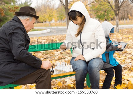 Attractive woman sitting on a park bench in the cold autumn weather playing chess with an elderly man while her young son amuses himself on a tablet computer behind her