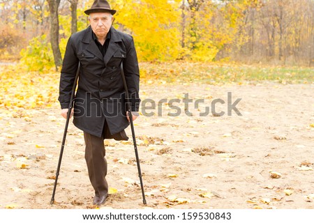 Lonely one-legged senior Caucasian man walking with crutches in an Autumn day in the park, with withered foliage in the background