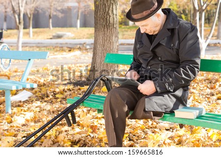 Retired handicapped man with one leg amputated sitting on a park bench in the autumn sun in an overcoat and hat using a tablet computer
