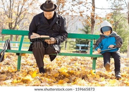 Grandfather and grandson amusing themselves reading together in the autumn sunshine sitting on a park bench with the old man reading a printed book and the child an e-book on his tablet-pc