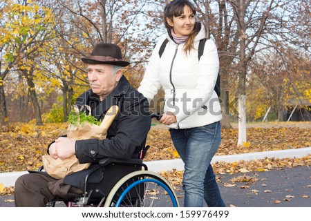 Smiling Female Carer Pushing An Old Handicapped Man In A Wheelchair Along The Street As They Return From Doing His Grocery Shopping