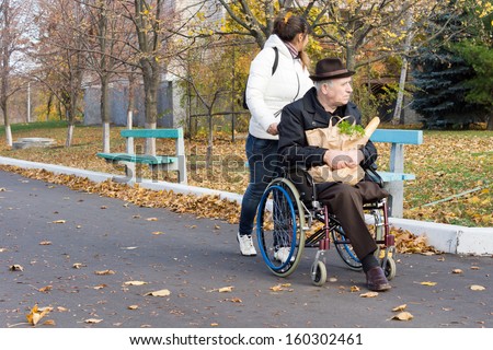 Female carer pushing a disabled man in a wheelchair with his groceries along the street turning to watch a young child playing in the park alongside