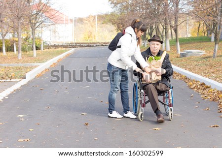 Daughter handing an elderly disabled man in a wheelchair a bag of groceries as she prepares to push him along the street after taking him shopping