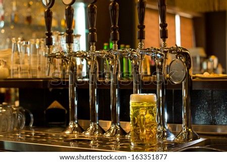 Large glass tankard filled with freshly poured golden draught beer poured from a row beer of stainless steel beer taps in a pub