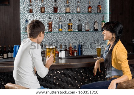 Young couple enjoying a date drinking a beer at the bar sitting at the counter chatting with a display of alcohol bottles on shelves behind them
