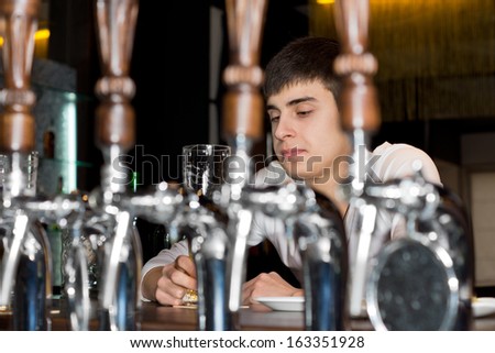 View between the stainless steel beer taps behind the counter of a young man seated alone at a bar drinking