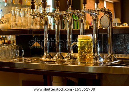 Row of stainless steel beer taps on a wooden counter for dispensing draught beer in a pub with a large glass tankard of beer alongside