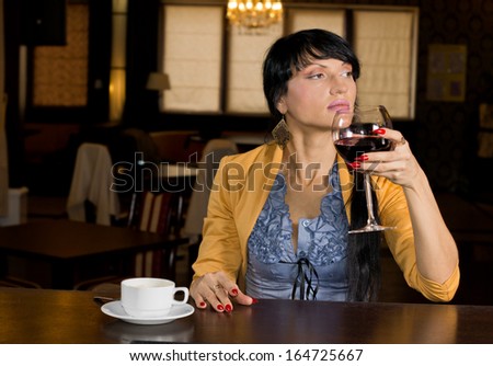 Young woman enjoying a large glass of red wine and cup of espresso coffee while sitting relaxing at a bar counter in a hotel or nightclub looking off to the side
