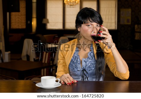 Attractive young woman drinking wine and coffee at a bar counter sipping from a large wineglass while glancing across at the camera