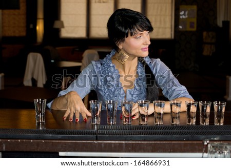 Woman with a row of vodka shot glasses lined up in front of her sitting at a bar counter smiling and looking away to the side