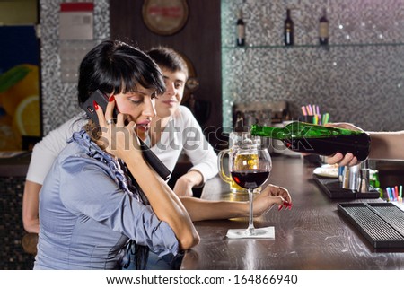 Woman chatting on her mobile while drinking at the bar in a nightclub listening to the conversation while the barman pours her another drink