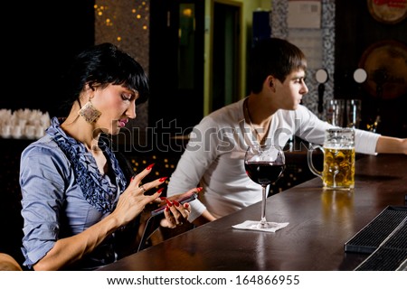 Stylish young woman sitting at the counter at the bar sending an sms message on her mobile phone, side view