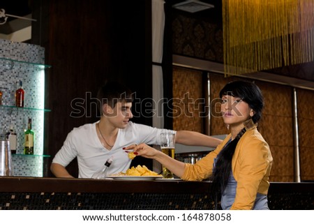 Beautiful stylish young Hispanic woman with long braided hair sitting at a bar counter drinking a pint of beer and eating snacks as the barman works behind the counter