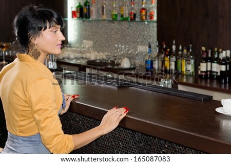 Young woman waiting for service at the bar counter sitting with her hands resting on the wood as she watches the barman who is off frame