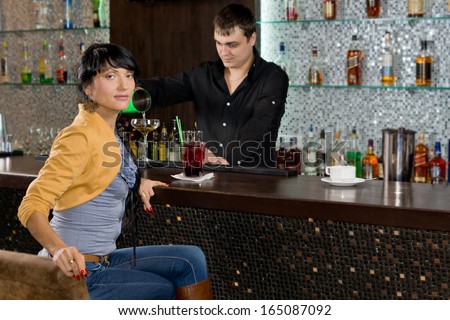Stylish woman sitting at the bar drinking alone while the barman works behind the counter mixing drinks for other customers