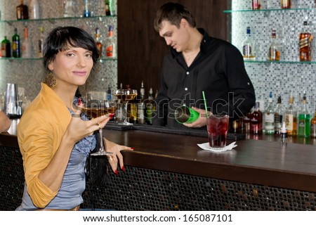 Beautiful young Hispanic woman drinking red wine at the bar turning to smile at the camera as the barman works behind the counter in the background