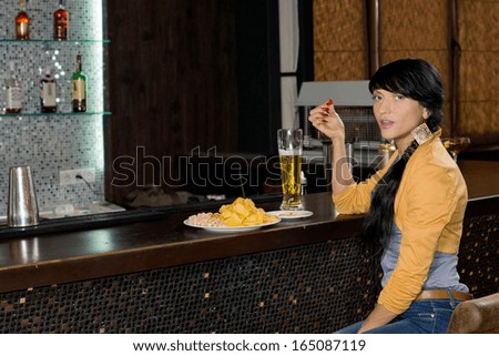 Stylish woman drinking alone at the bar counter enjoying a pint of draught beer and snacking on a plate of eats turning to look at the camera