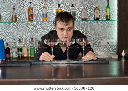 Bartender preparing three glasses of alcoholic cocktail to serve to customers bending down to check the levels of the liquid