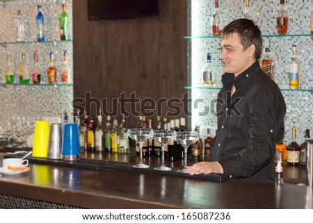 Smiling friendly barman serving alcoholic cocktails standing behind the bar counter with three cocktail glasses full of an exotic blend in front of him