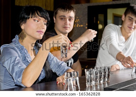 Young Hispanic woman drinking vodka shots at the counter in pub watched by her amused boyfriend and his friend
