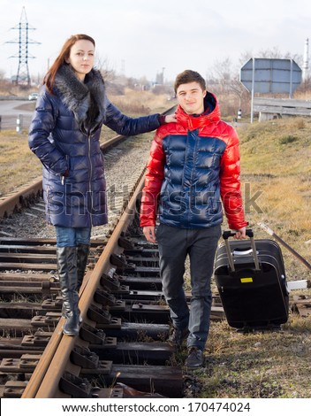 Young couple waiting for the train standing alongside the track with their packed suitcase with the young woman balanced on the metal track holding the mans shoulder