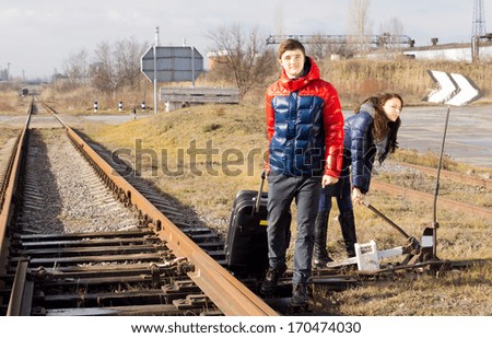 Young couple waiting for the train near a level crossing with a packed suitcase as the young woman fiddles with a lever alongside the track