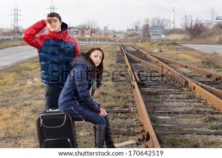 Young couple waiting for a train alongside a rural siding with the man peering into the distance watching and the woman relaxing sitting on their suitcase