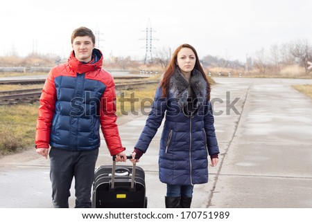 Smiling young couple pulling along a suitcase both holding onto the handle as they walk along a rural road alongside a railway line together