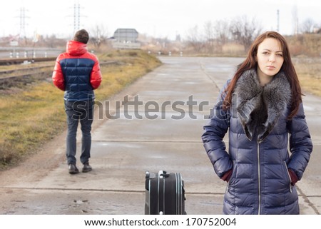 Young woman waiting with her suitcase for a lift in the middle of the road while a young man walks past in the opposite direction