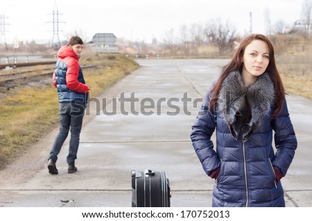 Attractive young woman waiting for a lift on a rural road with a packed suitcase being ogled by a young man as he strolls past