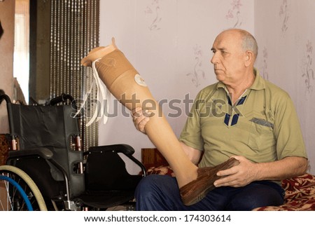 Elderly amputee sitting on his bed alongside his wheelchair holding an artificial leg in his hand as he prepares to fit it to his stump