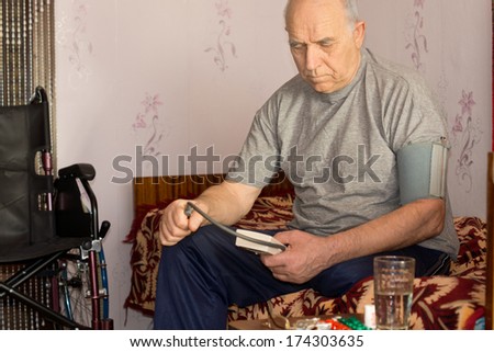 Senior disabled man taking his own blood pressure sitting on his bed alongside his wheelchair using a pressure cuff and sphygmomanometer
